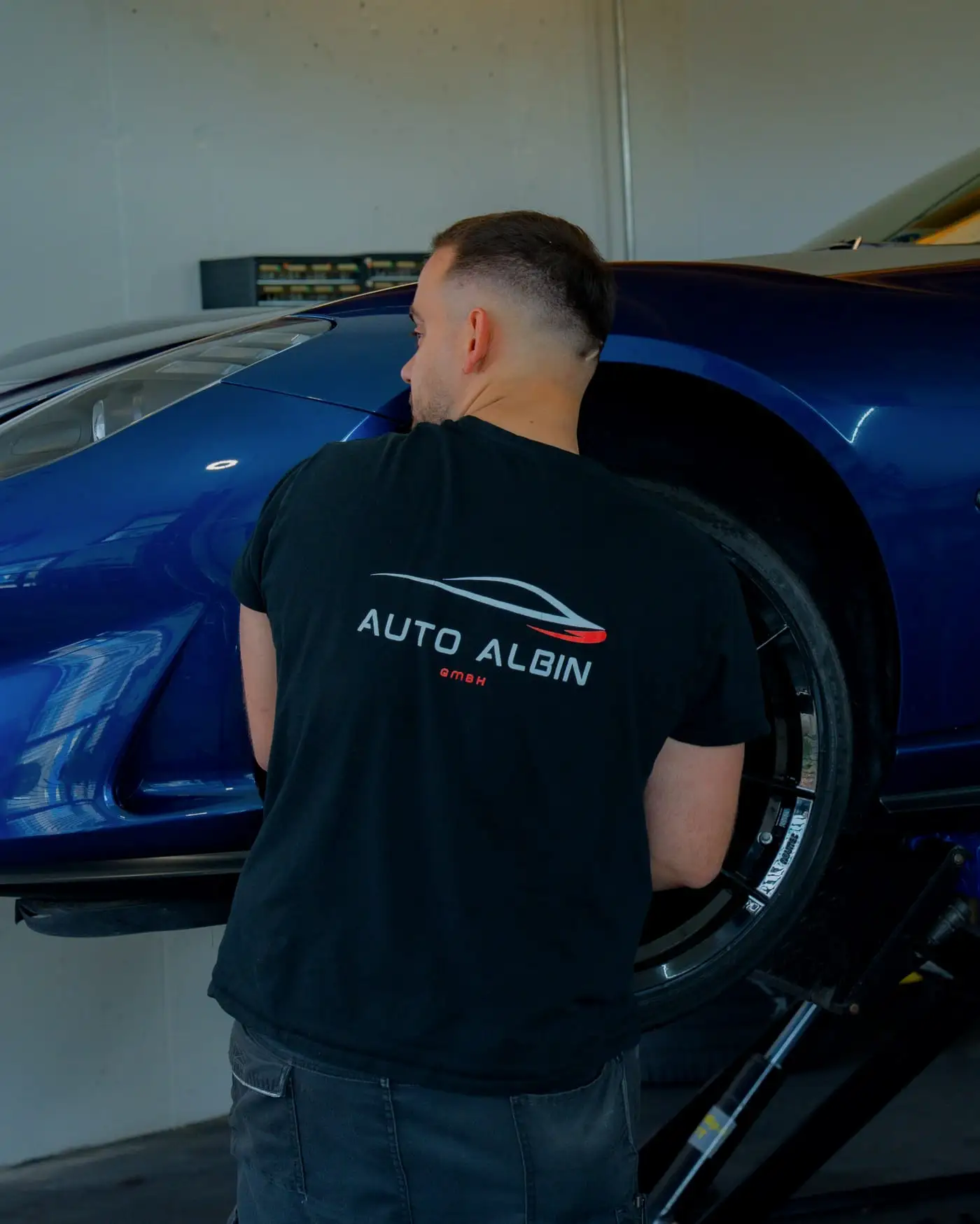 Mechaniker im Auto-Albin-Shirt montiert ein Rad an einem blauen Sportwagen auf der Hebebühne