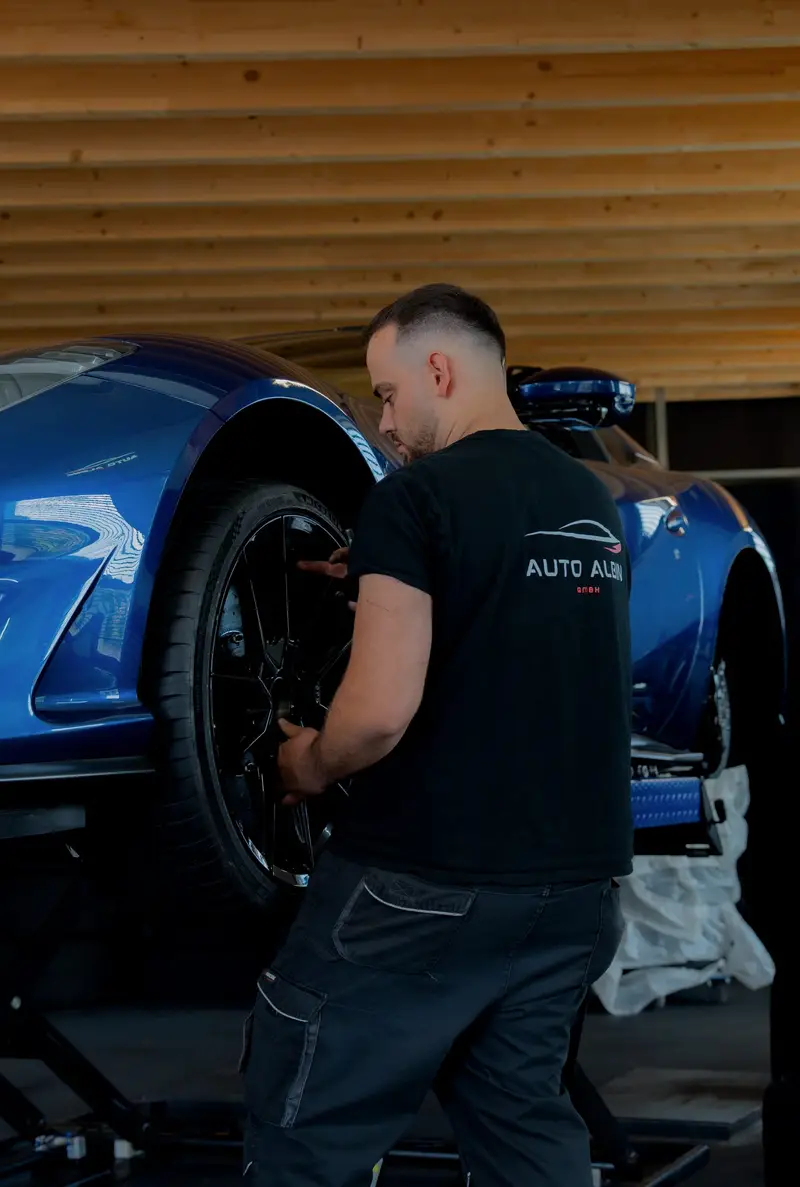 Mechaniker im Auto-Albin-Shirt montiert ein Rad an einem blauen Sportwagen auf der Hebebühne, unter der Holzdecke der Werkstatt in Sins AG