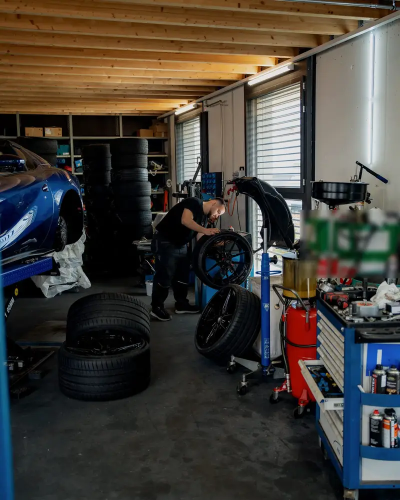 Werkstatthalle von Auto Albin unter der Holzdecke: Mechaniker an einem Rad, Reifen, Werkzeugwagen und ein Sportwagen auf der Hebebühne